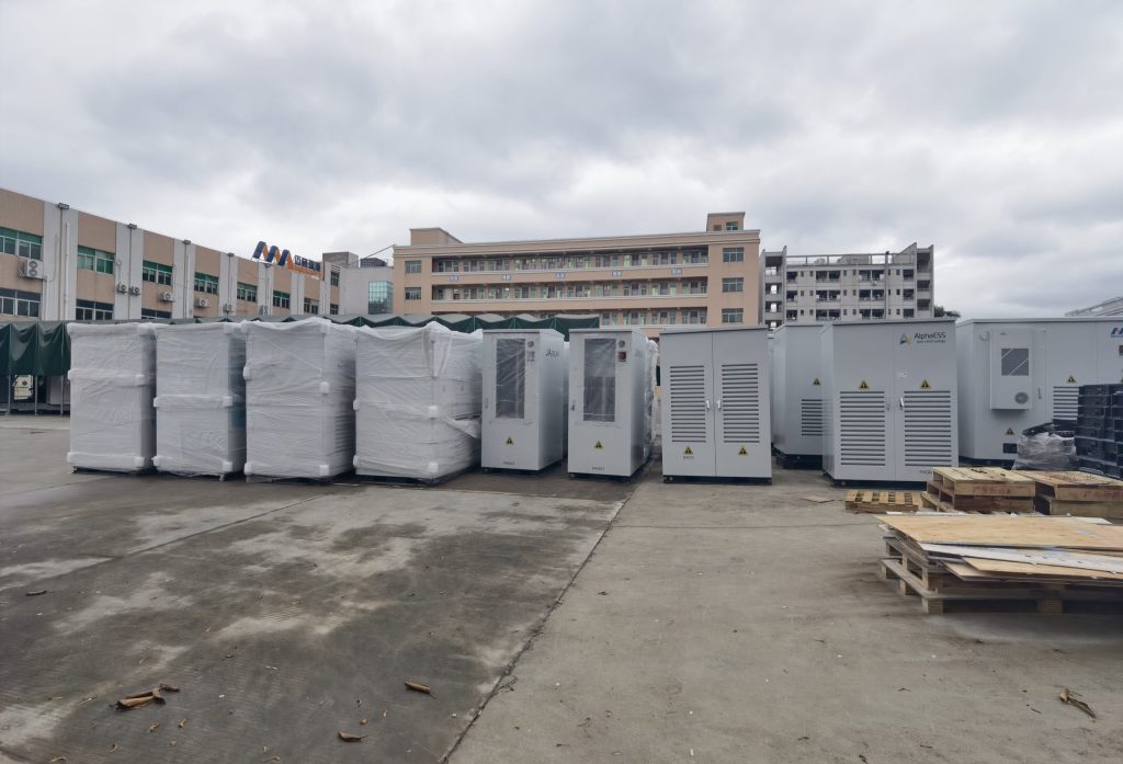 A wide-angle exterior photograph of a large collection of industrial battery energy storage systems (BESS), some plastic-wrapped for shipment and some unwrapped, lined up in a factory yard next to a multi-story manufacturing building. This showcases a fleet of MegSolid commercial and industrial solid-state energy storage cabinets.