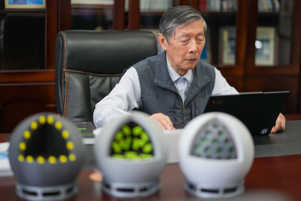 Elderly Chinese scientist Academician Chen Liquan wearing a grey vest, looking at a black laptop on a wooden desk. Three blurred crystal structure models sit in the foreground.