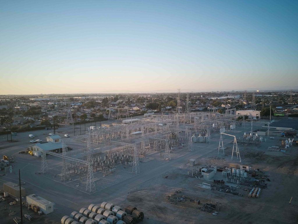 High-altitude drone photo of a large electrical substation site in Fresno during golden hour, showing high-voltage switchyards, transmission towers, and cable reels prepared for a MegSolid BESS installation.