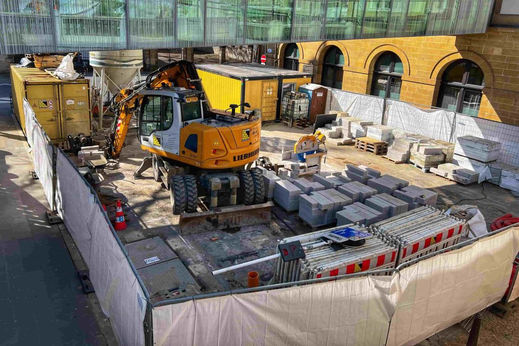 Elevated view of a fenced construction work zone in Munich, featuring a wheeled excavator, yellow utility containers, and stacked concrete paving blocks prepared for the installation of a MegSolid energy storage system.