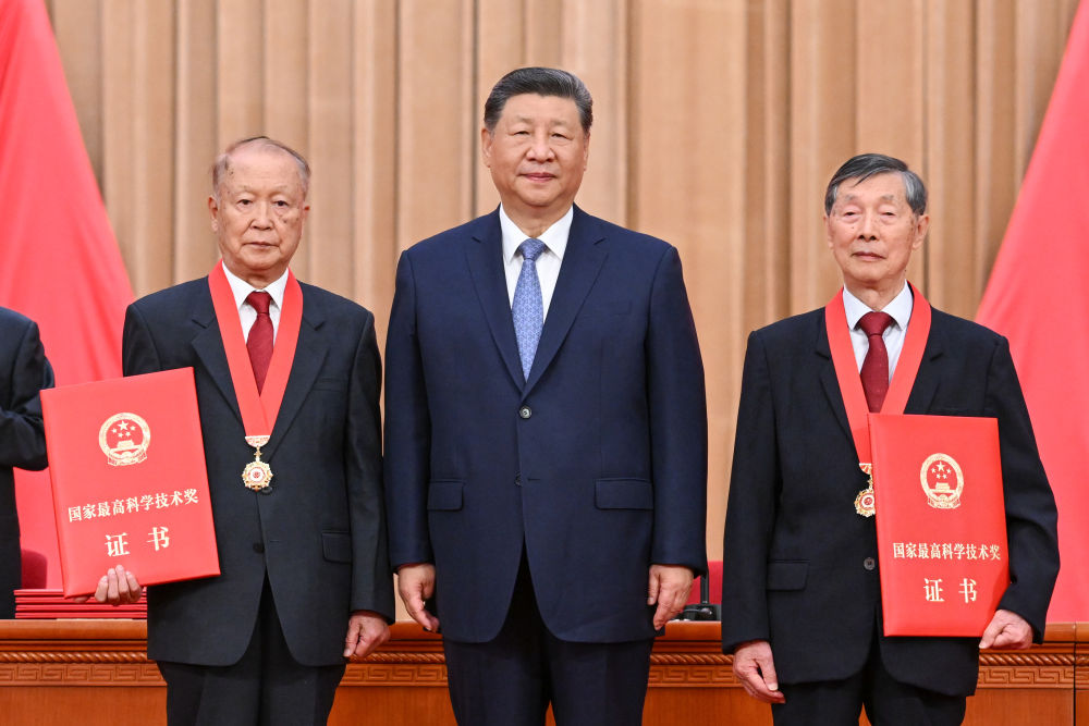 Chinese President Xi Jinping standing in the center with two elderly male scientists. The scientists are wearing dark suits, red ties, and gold medals, and each is holding a large red certificate that reads "State Preeminent Science and Technology Award Certificate" in Chinese.